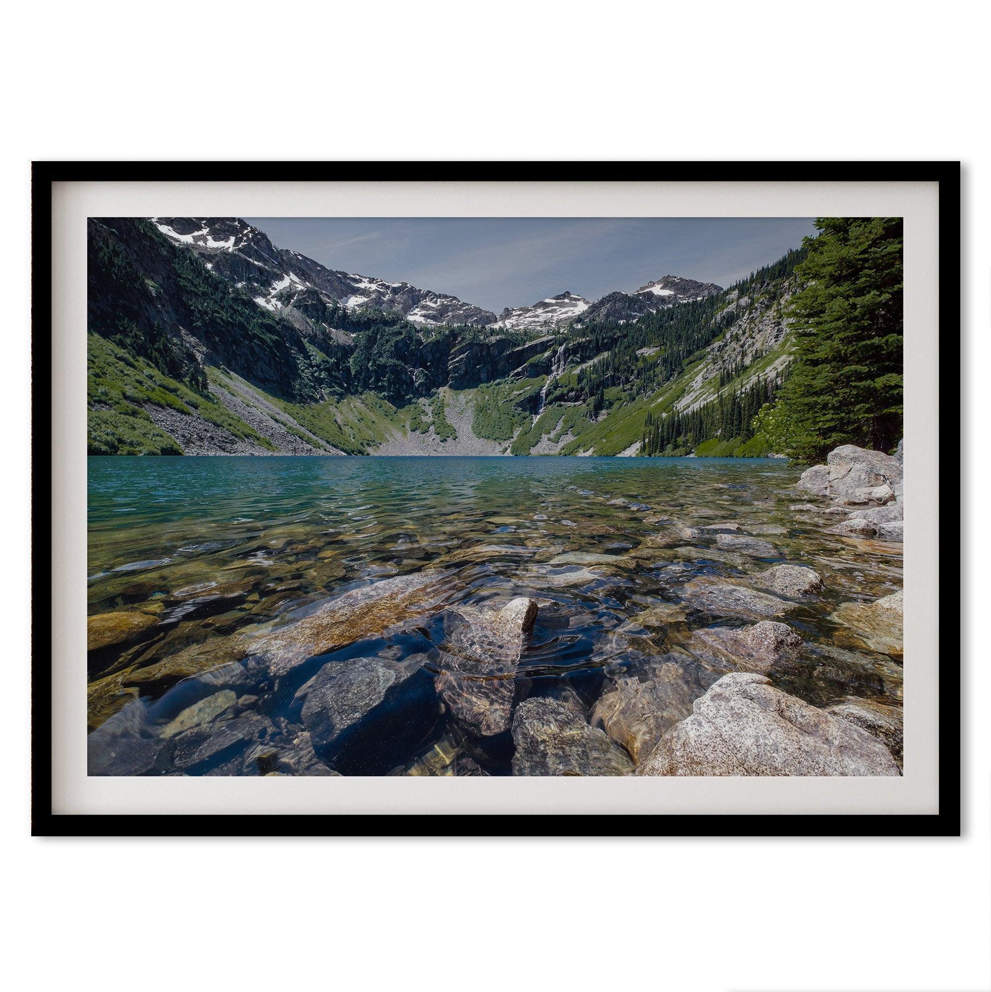 A fine art nature landscape framed print of blue lake in Washington State. The serene lake has vivid colors and clear water. So clear that you can see the stunning rocks at the bottom of the lake. In the backdrop, there are snow-covered mountains.