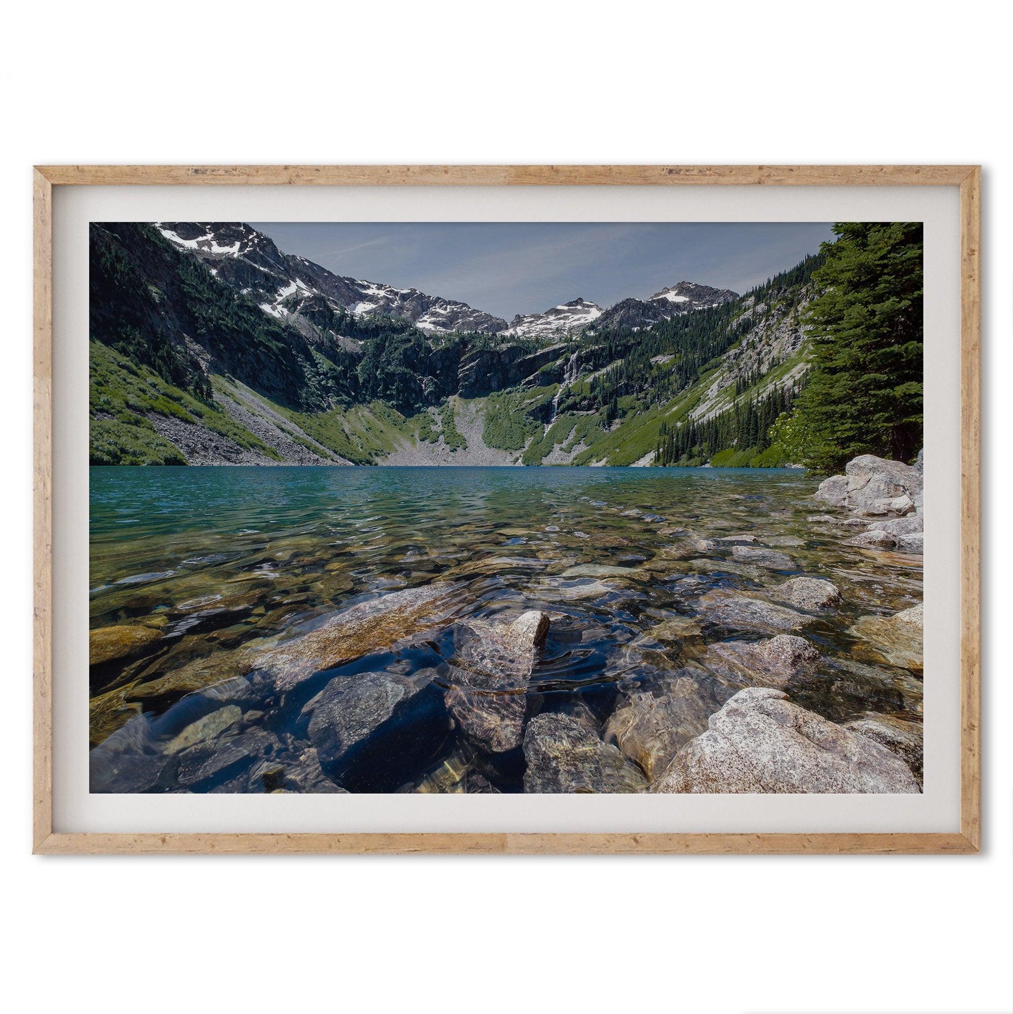 A fine art nature landscape framed print of blue lake in Washington State. The serene lake has vivid colors and clear water. So clear that you can see the stunning rocks at the bottom of the lake. In the backdrop, there are snow-covered mountains.