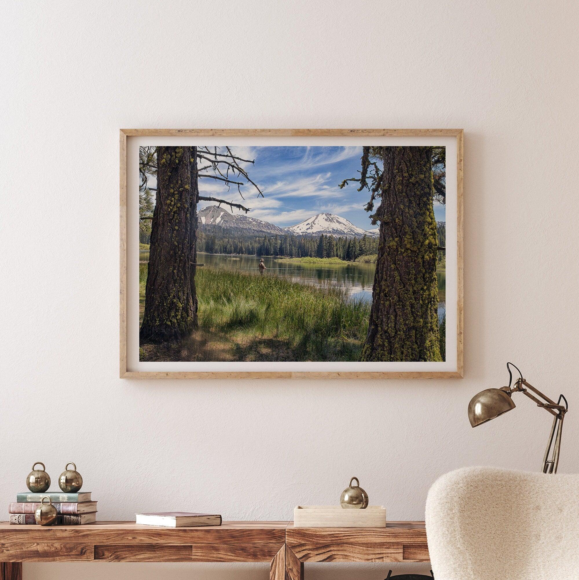 A framed or unframed mountain photo print capturing a fisherman fly fishing in a serene lake, with snow-capped mountains in the backdrop.