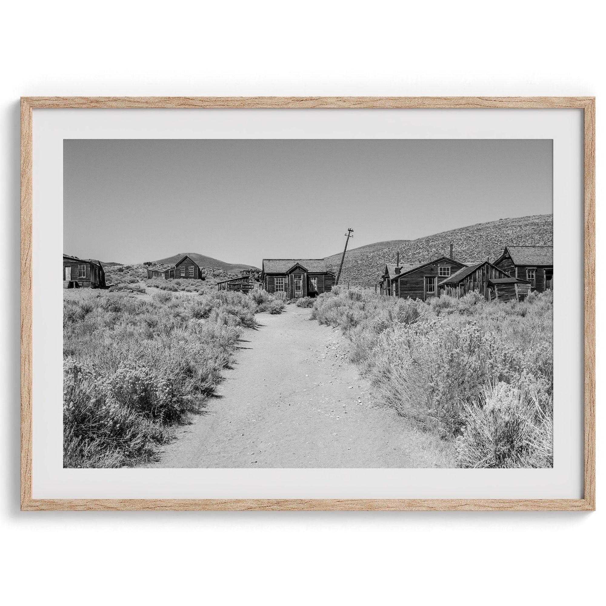 A fine art black and white desert print of a gold rush era town with a road and flowers leading to it. Taken in Bodie State Park, California.