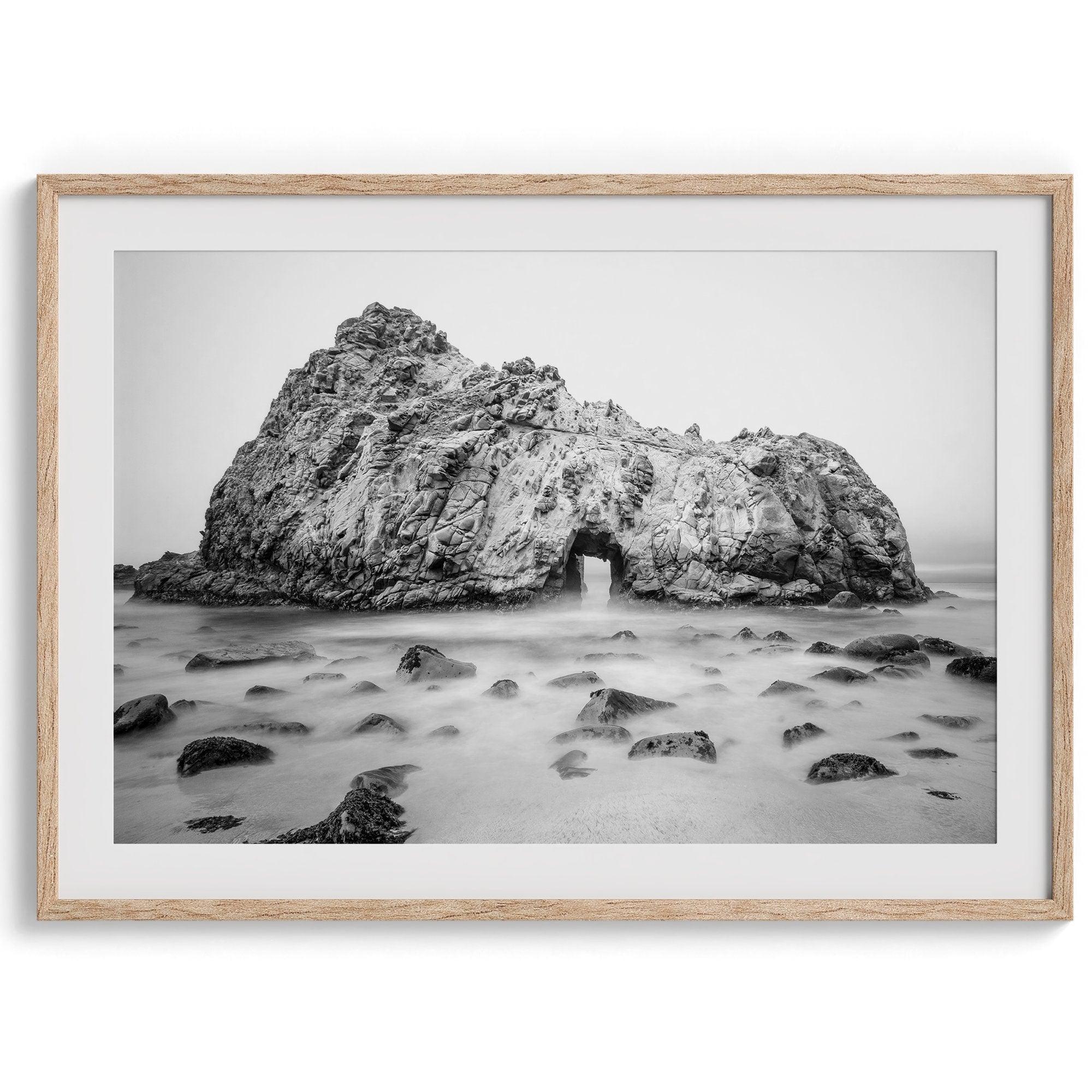 A fine art black and white print of Big Sur Pfeiffer Beach. This beach wall art shows a huge rock on the beach with an opening towards the open sea. This fine art photograph is shot in long exposure so the waves look creamy, gliding over the rocks