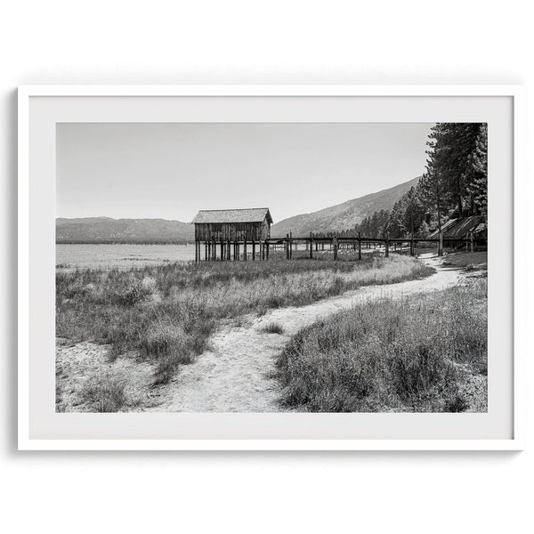 A fine art black and white vintage style framed photography print featuring a rustic old boathouse nested on a Lake Tahoe beach.