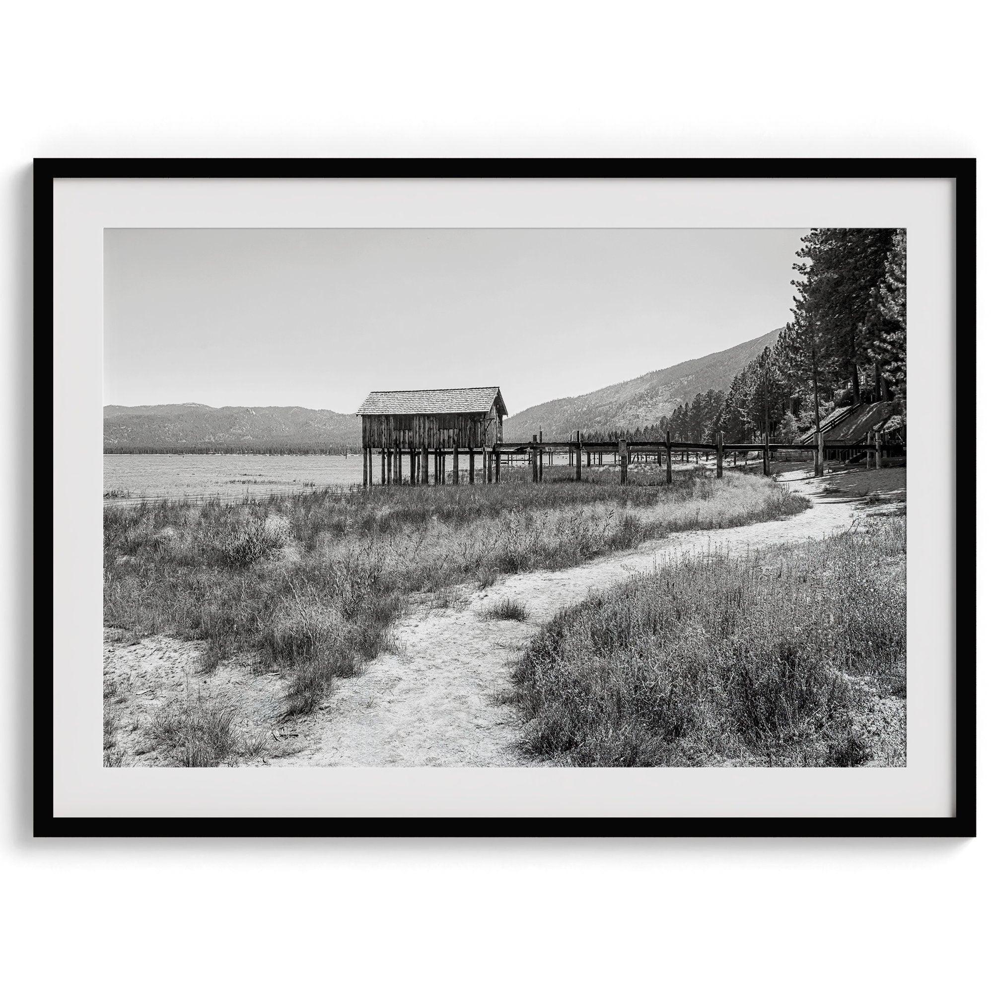 A fine art black and white vintage style framed photography print featuring a rustic old boathouse nested on a Lake Tahoe beach.