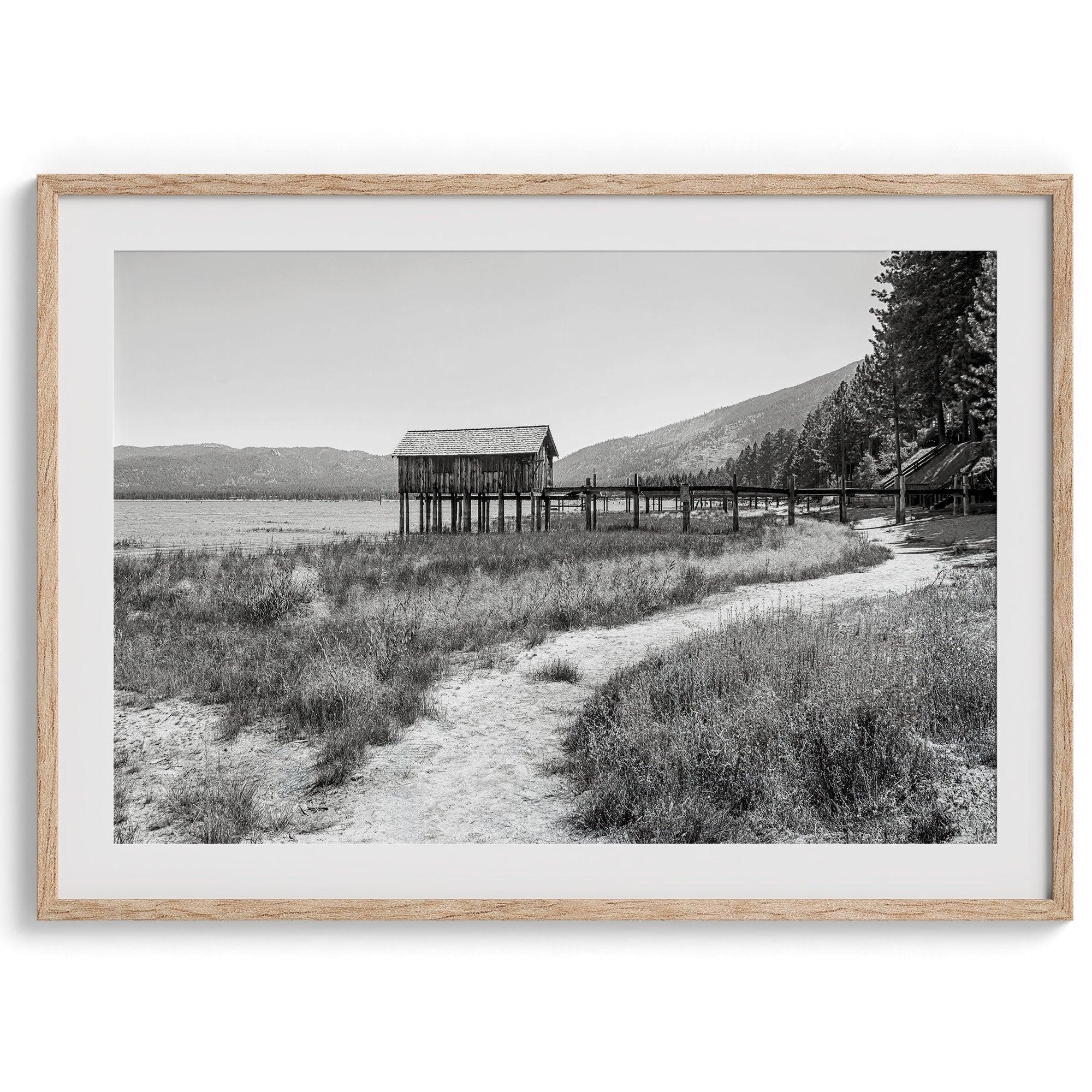 A fine art black and white vintage style framed photography print featuring a rustic old boathouse nested on a Lake Tahoe beach.