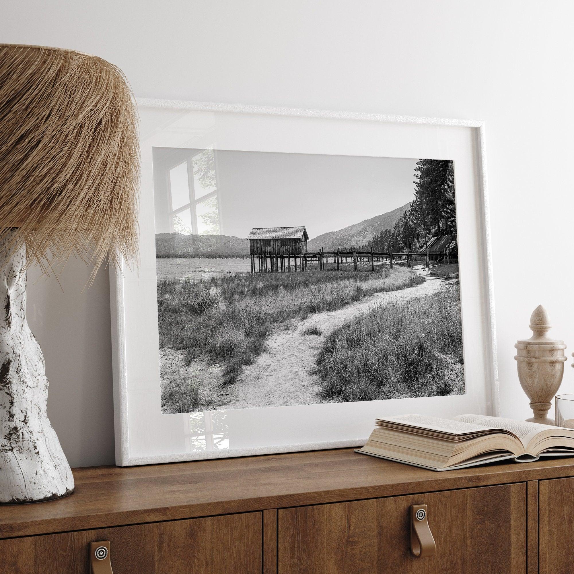 A fine art black and white vintage style framed photography print featuring a rustic old boathouse nested on a Lake Tahoe beach.