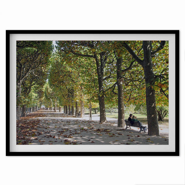 A fine art Paris print of a Parisian garden in fall showcasing a couple sitting on a bench under the fall color trees with foliage all around.