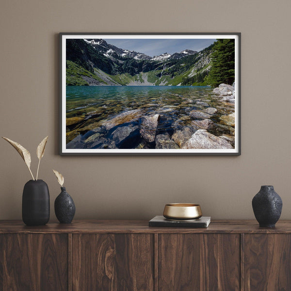 A fine art nature landscape framed print of blue lake in Washington State. The serene lake has vivid colors and clear water. So clear that you can see the stunning rocks at the bottom of the lake. In the backdrop, there are snow-covered mountains.