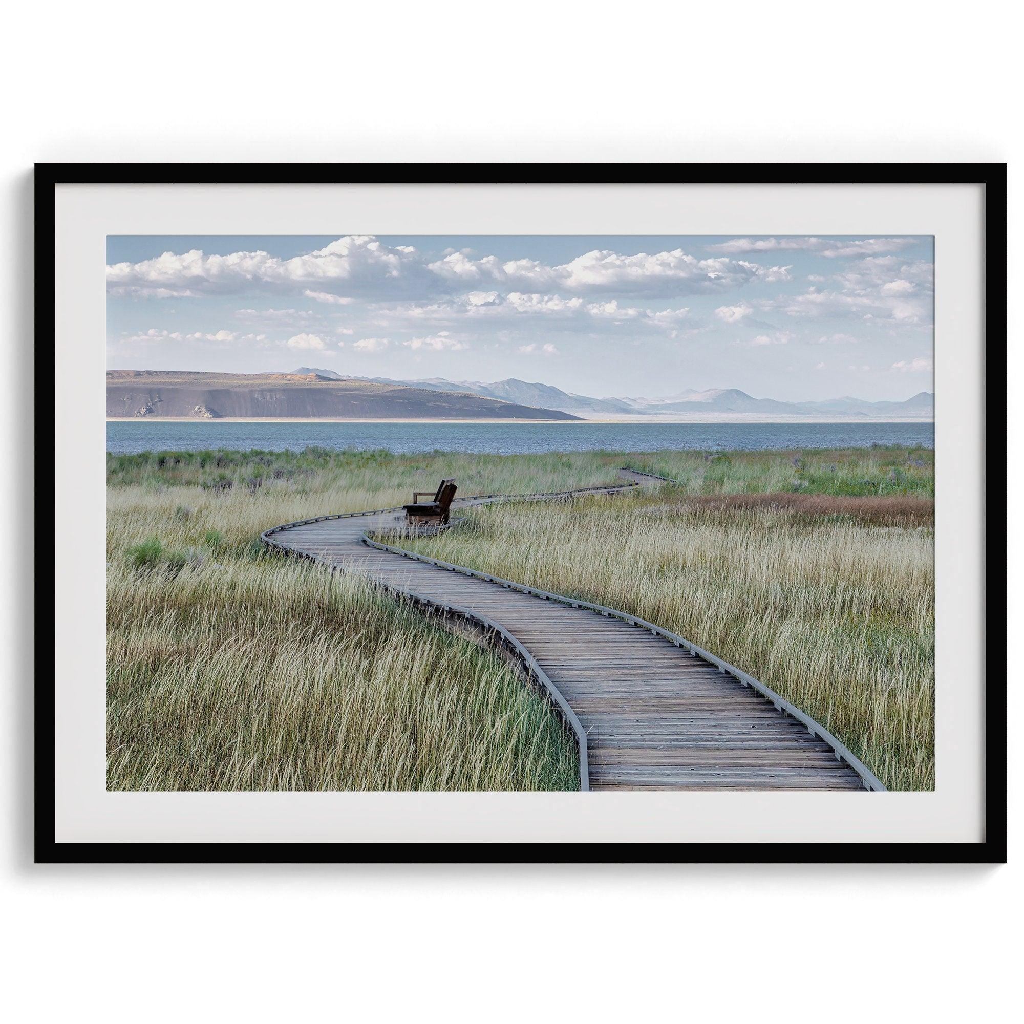 A fine art nature photography print featuring an inspiring winding path through the grassy wilderness of the eastern Sierra Nevada mountains toward Mono Lake. you can see the blue water of the lake in the background and a bench on the road.