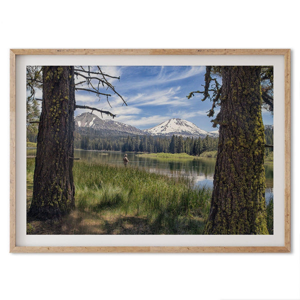 A framed or unframed mountain photo print capturing a fisherman fly fishing in a serene lake, with snow-capped mountains in the backdrop.