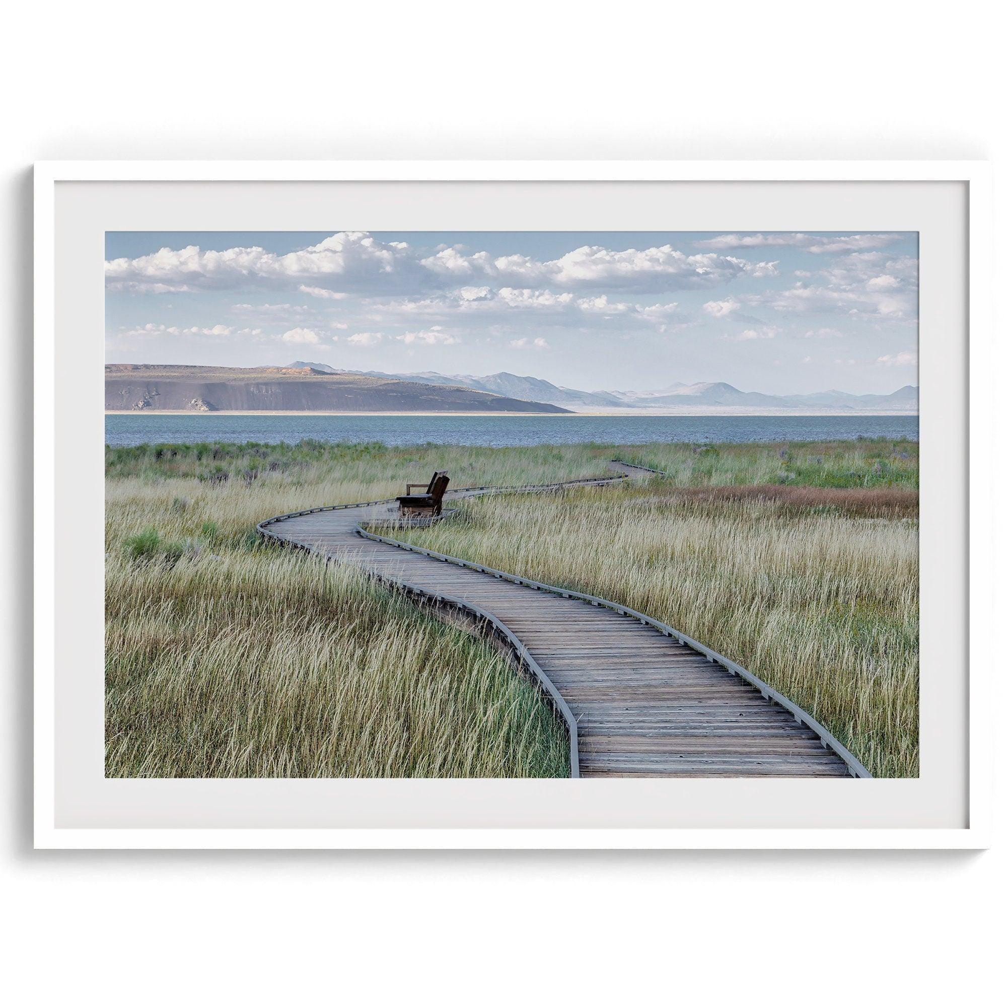 A fine art nature photography print featuring an inspiring winding path through the grassy wilderness of the eastern Sierra Nevada mountains toward Mono Lake. you can see the blue water of the lake in the background and a bench on the road.