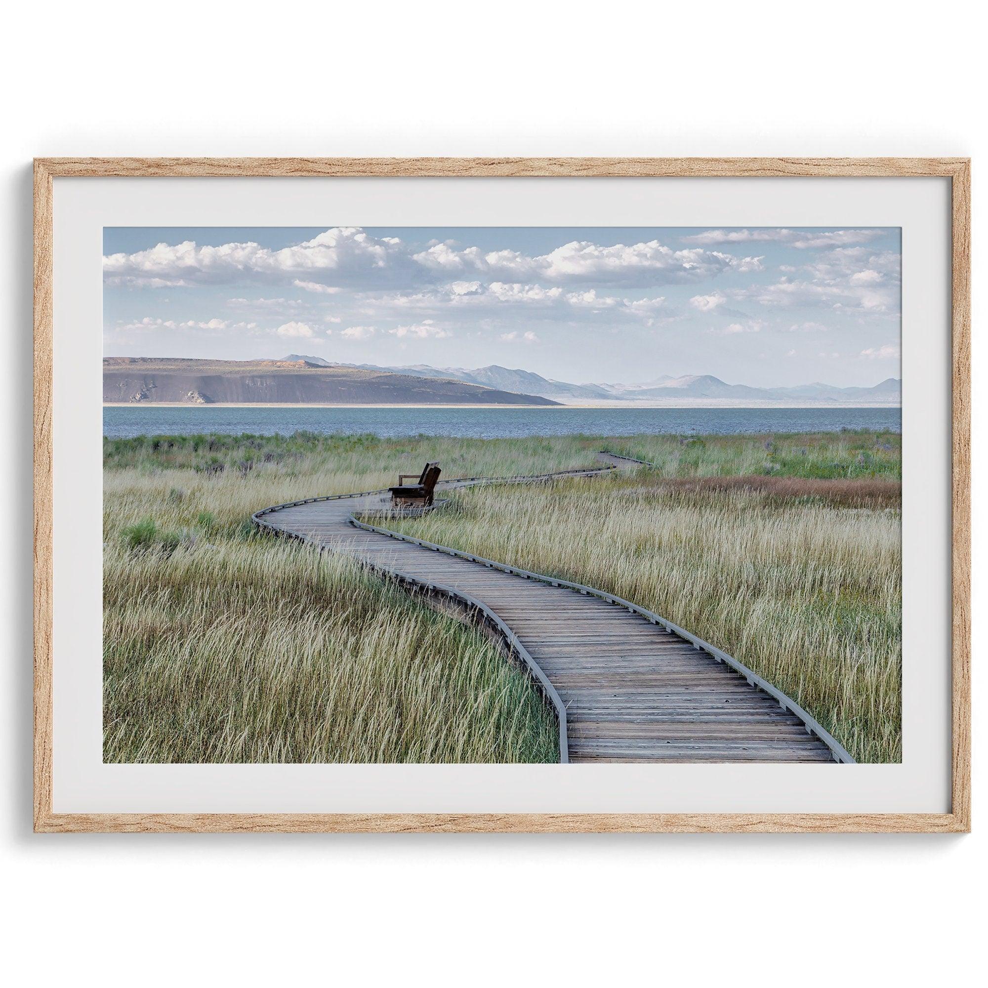 A fine art nature photography print featuring an inspiring winding path through the grassy wilderness of the eastern Sierra Nevada mountains toward Mono Lake. you can see the blue water of the lake in the background and a bench on the road.