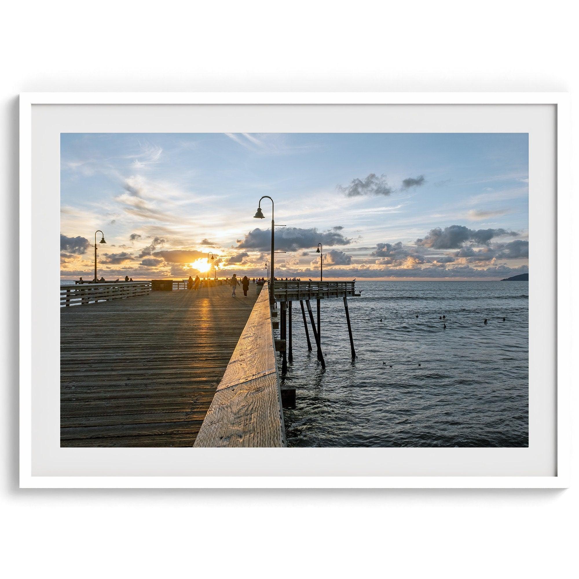 A fine art ocean sunset print featuring Pismo Beach Pier in Sunset with the surfers in the ocean and people enjoying a stroll on the pier.