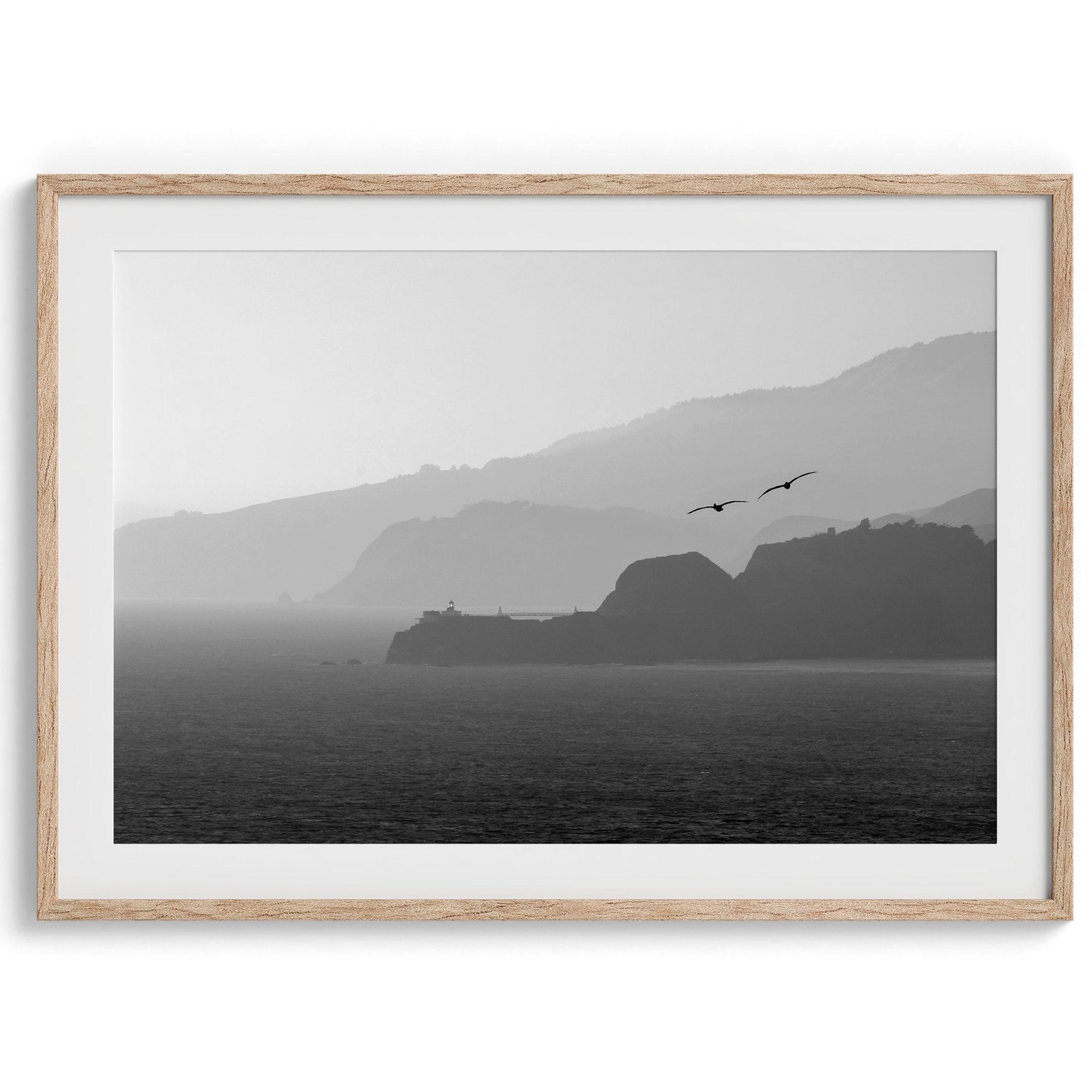 A black and white coastal wall art that showcases two Pelicans flying over the Pacific Ocean near the Golden Gate Bridge toward the layered mountains in the sunset.