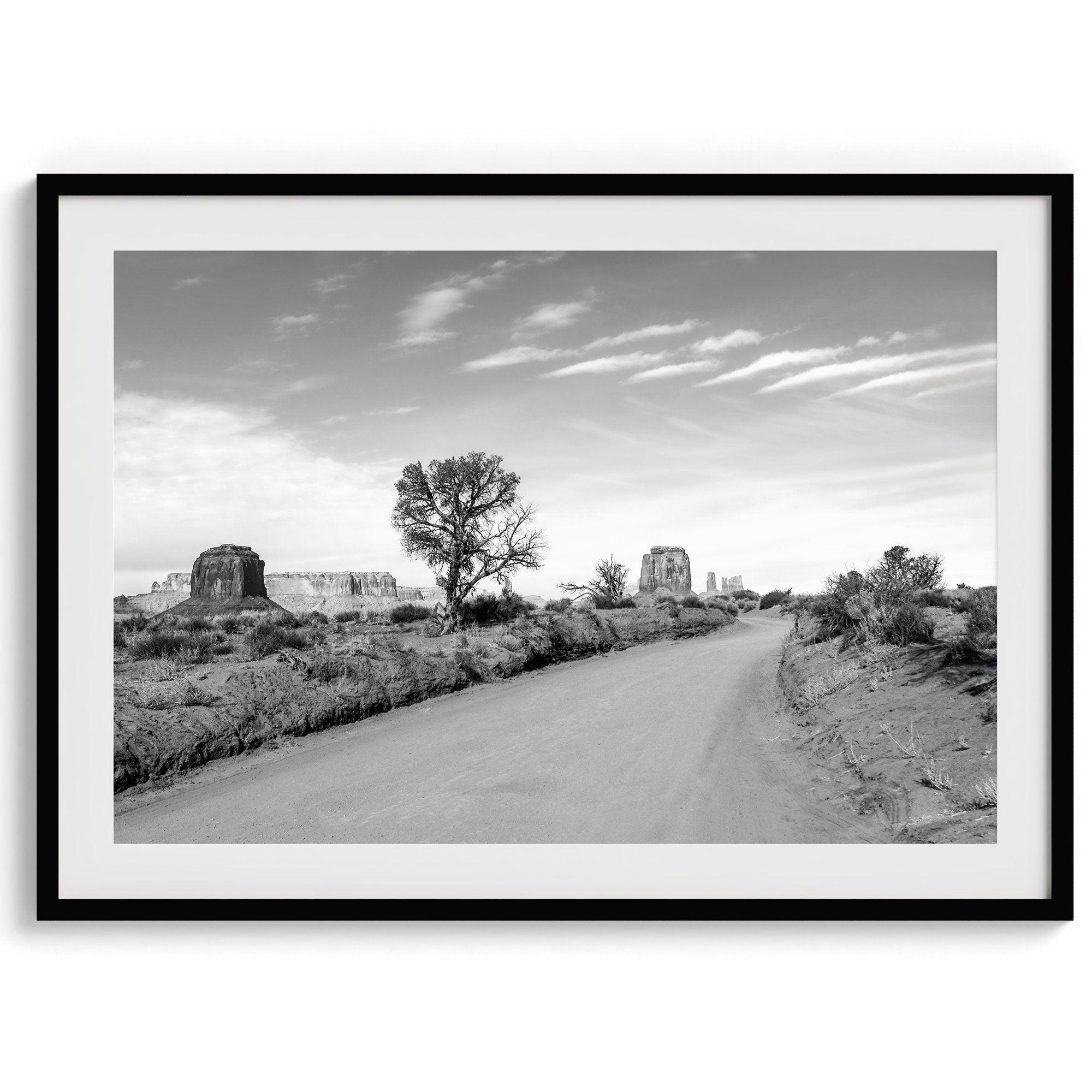 A fine black and white desert print, featuring Monument Valley in the Utah/ Arizona desert. The print shows a road that goes through the towering rocks and unique trees of monument valley.