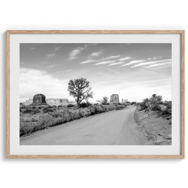 A fine black and white desert print, featuring Monument Valley in the Utah/ Arizona desert. The print shows a road that goes through the towering rocks and unique trees of monument valley.