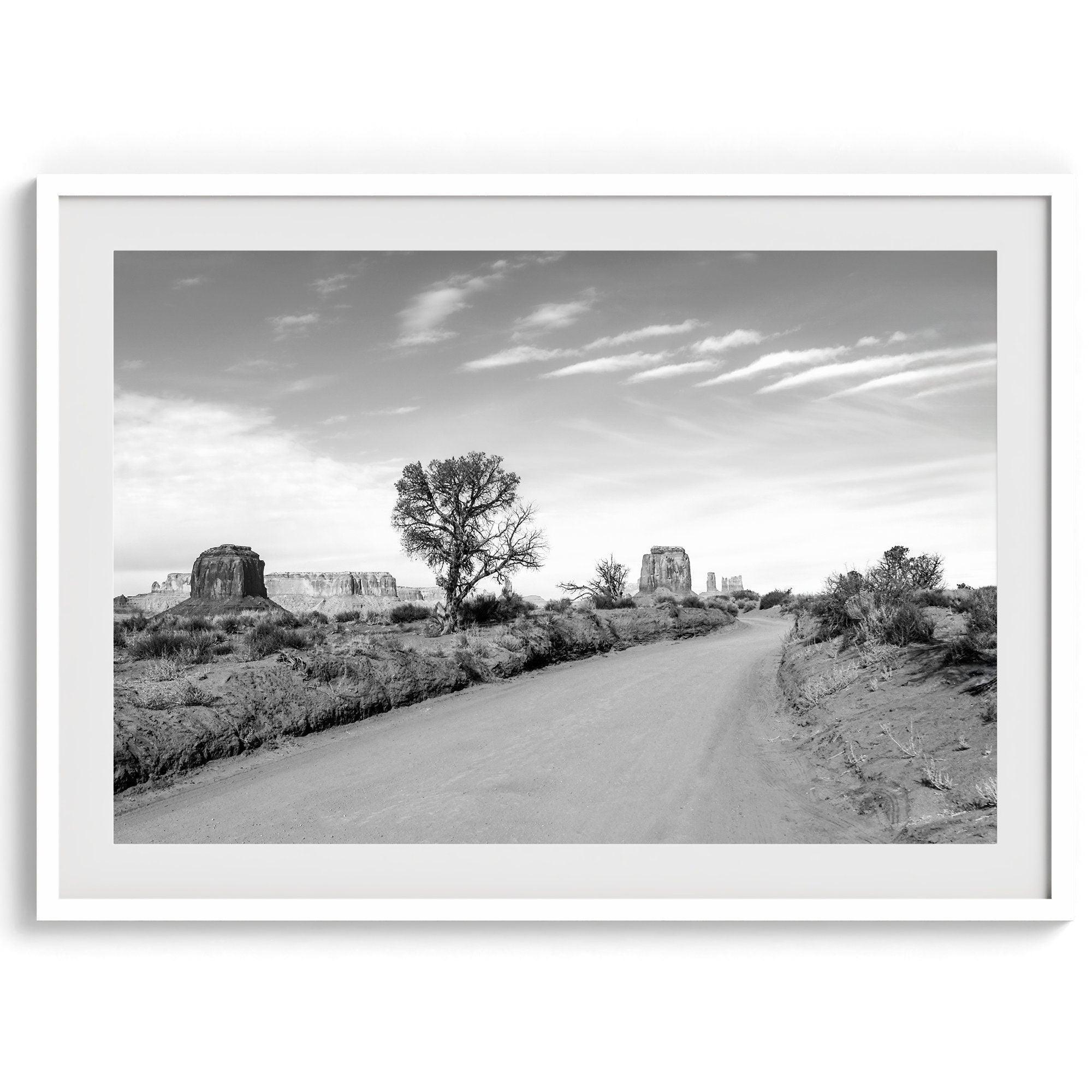 A fine black and white desert print, featuring Monument Valley in the Utah/ Arizona desert. The print shows a road that goes through the towering rocks and unique trees of monument valley.