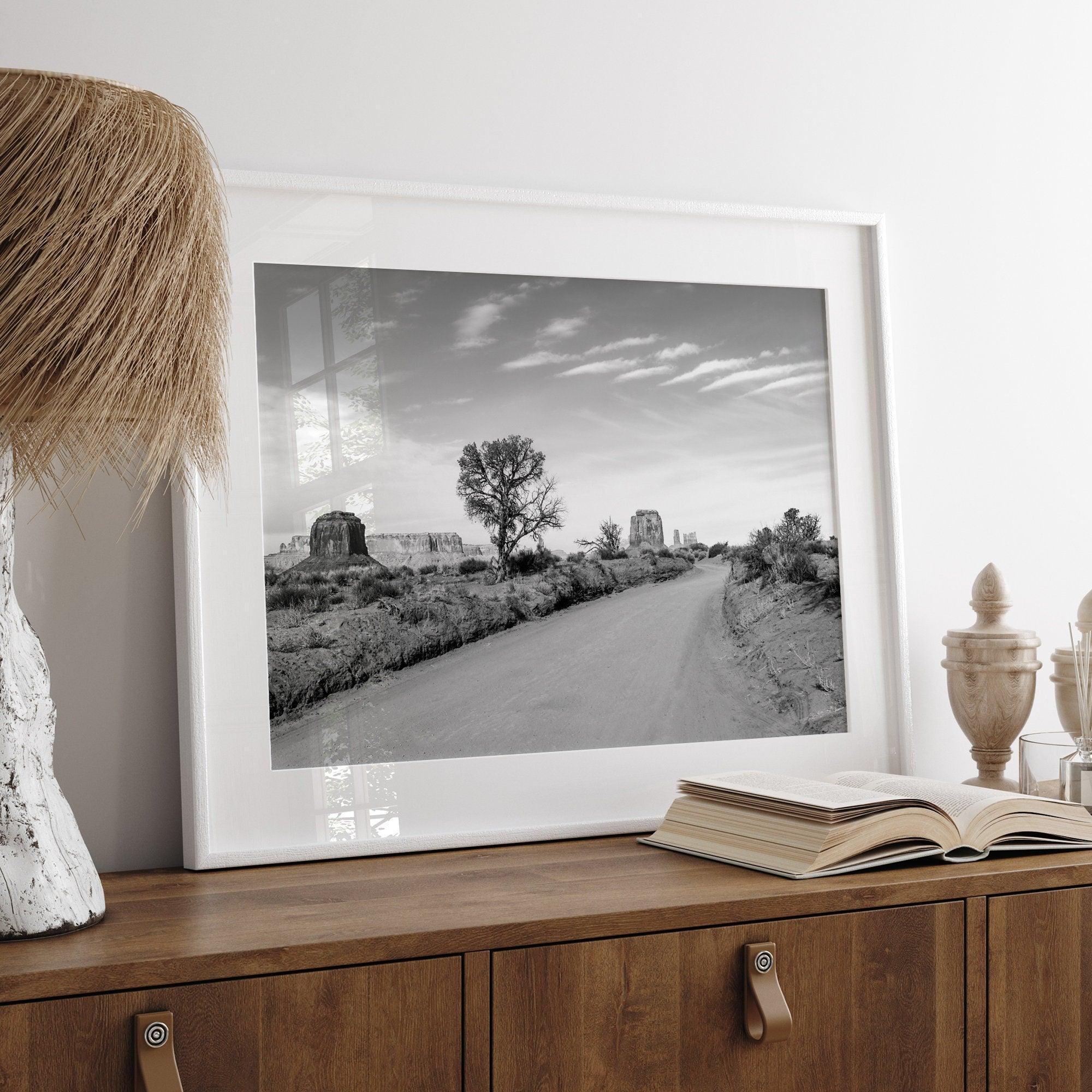 A fine black and white desert print, featuring Monument Valley in the Utah/ Arizona desert. The print shows a road that goes through the towering rocks and unique trees of monument valley.